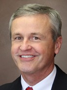 A man with short gray hair, wearing a black suit and red tie, smiling against a plain brown background. A man with short gray hair, wearing a black suit and red tie, smiling against a plain brown background.