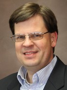 A man with glasses, short brown hair, and a slight smile, wearing a light blue plaid shirt and a dark blazer, against a plain brown background.