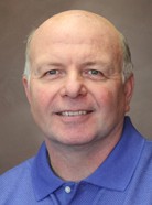 A bald man with a light complexion is wearing a blue collared shirt and smiling. The background is a plain, neutral brown.