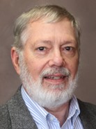 A man with short gray hair and a beard, wearing a gray sports coat over a light blue dress shirt, standing against a plain brown background.