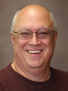 A man with glasses, short gray hair, and a brown sweater is smiling in front of a plain, brown background.