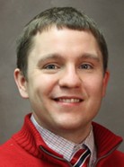 A man with short brown hair, wearing a red sweater and checkered shirt with a striped tie, smiles at the camera.