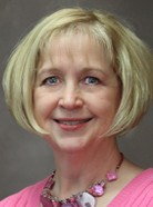 A woman with short blonde hair and blue eyes, wearing a pink top and a necklace with pink and purple beads, smiles at the camera.