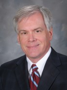 A middle-aged man in a suit and tie smiles slightly against a neutral gray background.
