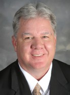 A man with short gray hair in a black suit, white shirt, and tie poses for a professional portrait.