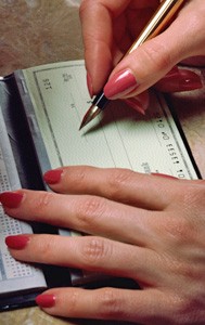 A person with red-painted nails writes on a check with a pen, using their left hand to steady the checkbook.