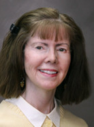 A woman with shoulder-length brown hair, wearing a light yellow top and a white collared shirt, smiles at the camera. She has a barrette in her hair and is set against a plain, dark background.