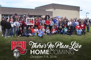 A large group of people, wearing a mix of casual and team apparel, gathered outdoors in front of a building. A banner reads "KCU There's No Place Like" at the bottom of the image, celebrating Kentucky Christian University alumni.