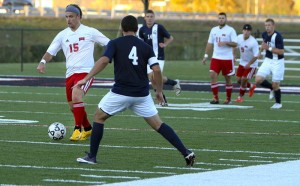 Soccer players from two teams compete for the ball on an outdoor field. One player in a dark blue jersey, possibly a Kentucky Christian University Alumni, focuses on an opponent in a white jersey with red shorts who is controlling the ball.