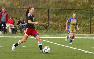 Two female soccer players are in action on a field. The player in red shorts, a Kentucky Christian University alumna, is controlling the ball, while the player in gold and blue follows behind. Several people are seated in the background.