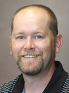 A man with a short beard and receding hairline is smiling. He is wearing a striped shirt with a black collar.