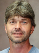 A man with short, light brown hair and a trimmed beard, wearing a blue shirt, smiles slightly in front of a plain background. A man with short, light brown hair and a trimmed beard, wearing a blue shirt, smiles slightly in front of a plain background.