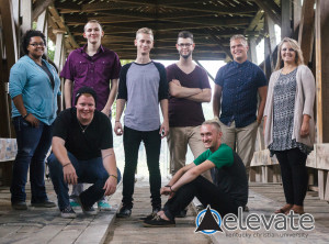 Group of eight people pose inside a wooden structure, with some standing and others crouching or sitting, dressed in casual clothing.