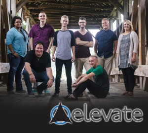 A group of eight people posing under a wooden structure. Four are standing at the back, while four are either sitting or crouching at the front. An overlay logo at the bottom reads, "Elevate.