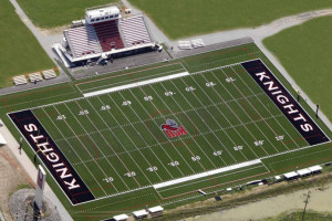 Aerial view of a football field with "Knights" written in the end zones and a knight logo at the center. A stadium with bleachers is visible on one side of the field, showcasing where future students can cheer on their team after successful admissions.