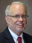 A man with glasses and a beard smiles, wearing a dark suit, white shirt, and red tie against a neutral background. A man with glasses and a beard smiles, wearing a dark suit, white shirt, and red tie against a neutral background.