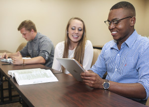 Three students sit at a table in a classroom. Two are smiling as they look at a tablet, while the third reads a book.