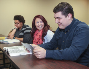 Three people are seated at a table. One person is reading a book, another is smiling, and a third person is using a tablet.