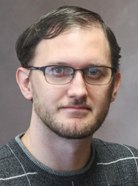 A man with short brown hair, glasses, and a beard poses for a portrait against a neutral background. He is wearing a gray sweater with white stripes.