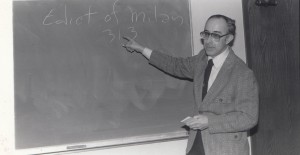 A man wearing a suit and tie is pointing to the words "Edict of Milan 313" written on a chalkboard. He is holding a piece of chalk and standing in front of the chalkboard.