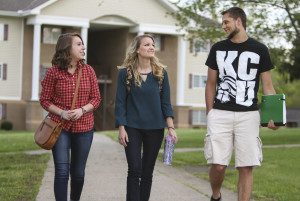 Three young adults walking outside, one wearing a red plaid shirt, another in a teal top, and the third in a black T-shirt and shorts, all appear to be conversing.