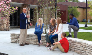 A teacher stands and speaks to five students sitting and kneeling on a stone bench outside a school building.